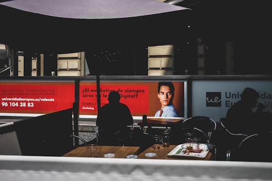 The image depicts a modern indoor setting with posters on a wall featuring a professional headshot and text related to marketing. Silhouetted figures are seated at tables in the foreground, casting shadows. The lighting is dim, creating contrast between the illuminated posters and darker seating area.