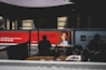 The image depicts a modern indoor setting with posters on a wall featuring a professional headshot and text related to marketing. Silhouetted figures are seated at tables in the foreground, casting shadows. The lighting is dim, creating contrast between the illuminated posters and darker seating area.