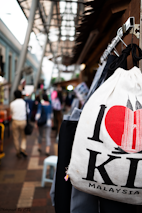 A bustling street scene with a focus on a white bag featuring a design of a red heart and the Petronas Towers, with the words 'I Love KL Malaysia'. People walking in the background are blurred, suggesting movement and a busy atmosphere. The setting appears to be an outdoor market with a mix of structures and overhead coverings.