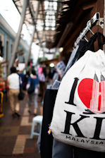 A bustling street scene with a focus on a white bag featuring a design of a red heart and the Petronas Towers, with the words 'I Love KL Malaysia'. People walking in the background are blurred, suggesting movement and a busy atmosphere. The setting appears to be an outdoor market with a mix of structures and overhead coverings.