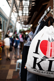 A bustling street scene with a focus on a white bag featuring a design of a red heart and the Petronas Towers, with the words 'I Love KL Malaysia'. People walking in the background are blurred, suggesting movement and a busy atmosphere. The setting appears to be an outdoor market with a mix of structures and overhead coverings.