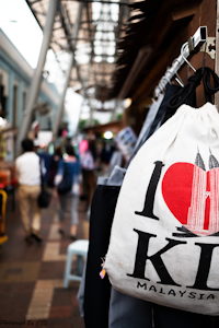 A bustling street scene with a focus on a white bag featuring a design of a red heart and the Petronas Towers, with the words 'I Love KL Malaysia'. People walking in the background are blurred, suggesting movement and a busy atmosphere. The setting appears to be an outdoor market with a mix of structures and overhead coverings.