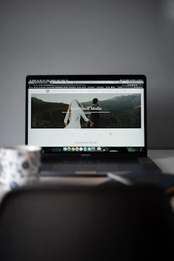 A laptop displaying a website with a large header image of a couple holding hands and looking towards a mountainous landscape. Text on the screen reads 'Randelhoff Media.' In the foreground, a blurred coffee cup with a floral pattern is visible.