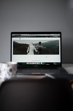 A laptop displaying a website with a large header image of a couple holding hands and looking towards a mountainous landscape. Text on the screen reads 'Randelhoff Media.' In the foreground, a blurred coffee cup with a floral pattern is visible.