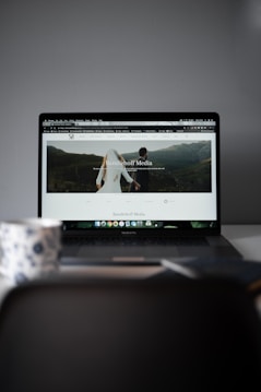 A laptop displaying a website with a large header image of a couple holding hands and looking towards a mountainous landscape. Text on the screen reads 'Randelhoff Media.' In the foreground, a blurred coffee cup with a floral pattern is visible.