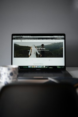 A laptop displaying a website with a large header image of a couple holding hands and looking towards a mountainous landscape. Text on the screen reads 'Randelhoff Media.' In the foreground, a blurred coffee cup with a floral pattern is visible.