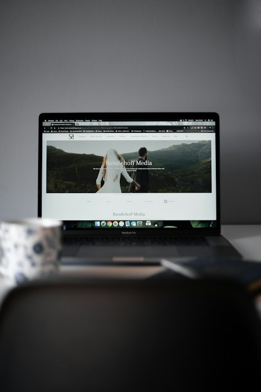 A laptop displaying a website with a large header image of a couple holding hands and looking towards a mountainous landscape. Text on the screen reads 'Randelhoff Media.' In the foreground, a blurred coffee cup with a floral pattern is visible.