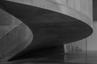 A sleek, modern architectural structure with curved, concrete elements that create a sense of fluidity. The design incorporates smooth surfaces and sharp edges, resulting in a minimalist, yet striking appearance.