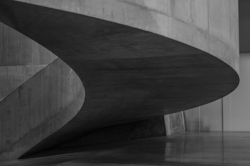 A sleek, modern architectural structure with curved, concrete elements that create a sense of fluidity. The design incorporates smooth surfaces and sharp edges, resulting in a minimalist, yet striking appearance.