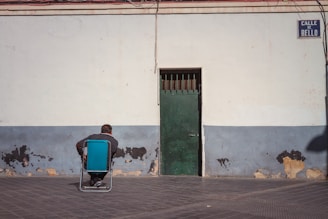 A person sits on a folding chair facing a wall with peeling paint. The wall has a faded blue base and a green door at the center. Overhead, a sign reads 'Calle de Bello.' There are visible wires hanging above.