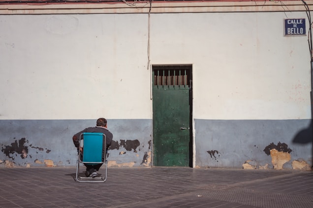 A person sits on a folding chair facing a wall with peeling paint. The wall has a faded blue base and a green door at the center. Overhead, a sign reads 'Calle de Bello.' There are visible wires hanging above.