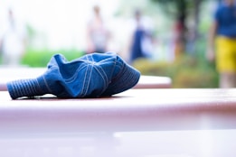 A blue denim cap is placed on a smooth, reflective surface, possibly a table or bench, outdoors. The background is blurred, suggesting a park or outdoor setting with people in the distance, creating a sense of depth.
