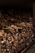 A large stack of chopped firewood is neatly arranged under a shelter. The logs vary in size, with exposed cut surfaces showing shades of brown and beige. Some pieces have remnants of bark and moss, indicating the natural origin of the wood. The setting suggests a rustic or outdoor environment.
