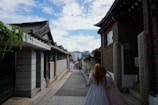 A narrow street flanked by traditional Korean houses with tiled roofs, leading to a view of modern city buildings in the background. Two people in traditional attire walk down the street, suggesting a blend of historical and contemporary elements.