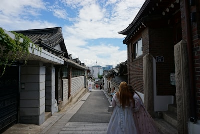 A narrow street flanked by traditional Korean houses with tiled roofs, leading to a view of modern city buildings in the background. Two people in traditional attire walk down the street, suggesting a blend of historical and contemporary elements.