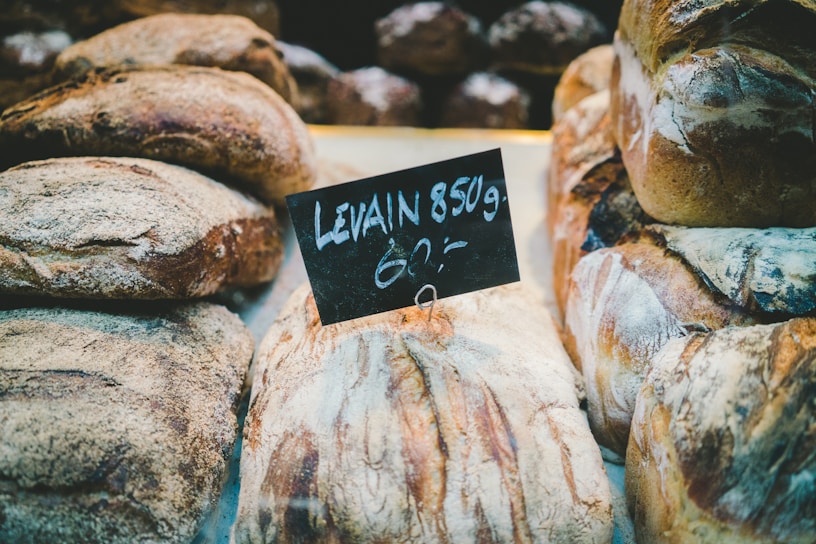 Loaves of crusty artisan bread are displayed on a counter with a small chalkboard sign indicating 'Levain 850g'. The breads have a rustic appearance with a dusting of flour and textured, golden-brown crusts. The setting suggests a bakery or market environment.
