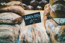 Loaves of crusty artisan bread are displayed on a counter with a small chalkboard sign indicating 'Levain 850g'. The breads have a rustic appearance with a dusting of flour and textured, golden-brown crusts. The setting suggests a bakery or market environment.