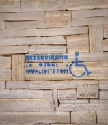 A stone wall with a stencil of a wheelchair symbol and text in blue indicating a reserved space for people with disabilities. Additionally, there is a simple drawing of a face and some text underneath in black.