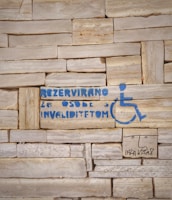 A stone wall with a stencil of a wheelchair symbol and text in blue indicating a reserved space for people with disabilities. Additionally, there is a simple drawing of a face and some text underneath in black.