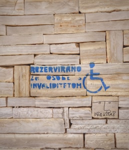 A stone wall with a stencil of a wheelchair symbol and text in blue indicating a reserved space for people with disabilities. Additionally, there is a simple drawing of a face and some text underneath in black.