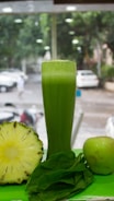 A tall glass filled with green juice is surrounded by a halved pineapple, a fresh apple, and a handful of spinach leaves. The background shows a blurred view of a street with parked cars and trees.