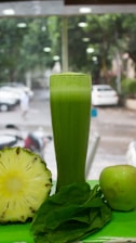 A tall glass filled with green juice is surrounded by a halved pineapple, a fresh apple, and a handful of spinach leaves. The background shows a blurred view of a street with parked cars and trees.