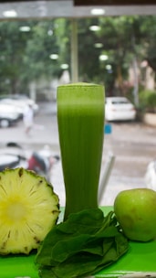 A tall glass filled with green juice is surrounded by a halved pineapple, a fresh apple, and a handful of spinach leaves. The background shows a blurred view of a street with parked cars and trees.