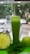 A tall glass filled with green juice is surrounded by a halved pineapple, a fresh apple, and a handful of spinach leaves. The background shows a blurred view of a street with parked cars and trees.