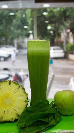 A tall glass filled with green juice is surrounded by a halved pineapple, a fresh apple, and a handful of spinach leaves. The background shows a blurred view of a street with parked cars and trees.