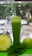 A tall glass filled with green juice is surrounded by a halved pineapple, a fresh apple, and a handful of spinach leaves. The background shows a blurred view of a street with parked cars and trees.
