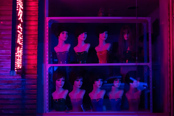 Mannequin heads with various hairstyles and painted red busts are displayed on shelves behind a glass window. Neon pink and blue lighting creates a dramatic and surreal atmosphere. An illuminated sign with indecipherable text is visible on the left side.