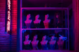 Mannequin heads with various hairstyles and painted red busts are displayed on shelves behind a glass window. Neon pink and blue lighting creates a dramatic and surreal atmosphere. An illuminated sign with indecipherable text is visible on the left side.