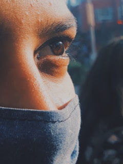 A close-up of a person's eye with strong lighting on the left side, wearing a dark face mask. The background is blurred, focusing attention on the eye and face.