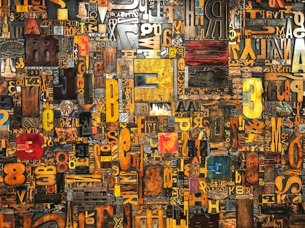 A vibrant and eclectic collage of various letters and numbers in different fonts, sizes, and colors arranged in a dense and intricate pattern. The elements appear as if they are made from different materials, including wood and metal, giving a textured and artistic feel.