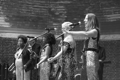 A group of four women are performing on stage with microphones. They are dressed in sparkling, sequined outfits and are singing passionately. The background includes a brick wall and a wooden structure above them. Additional musicians or performers are faintly visible in the background.