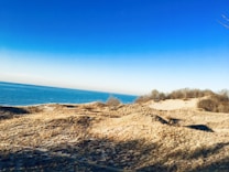 A serene landscape of sand dunes with sparse vegetation, set against the backdrop of a vast blue sky meeting a calm sea. The colors are vibrant with a clear gradient from pale to deep blue in the sky, and warm sandy tones.