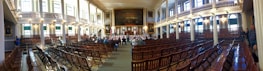 A large, historic meeting hall with rows of wooden chairs filling the room. High ceilings and tall windows allow natural light to flood the space. Ornate chandeliers hang from the ceiling, and an American flag decorates the stage. There are a few people scattered around the room, which features decorative paintings and intricate architectural details.