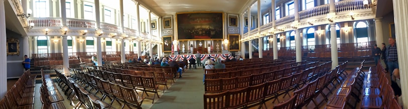 A large, historic meeting hall with rows of wooden chairs filling the room. High ceilings and tall windows allow natural light to flood the space. Ornate chandeliers hang from the ceiling, and an American flag decorates the stage. There are a few people scattered around the room, which features decorative paintings and intricate architectural details.