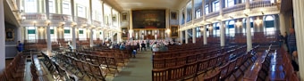 A large, historic meeting hall with rows of wooden chairs filling the room. High ceilings and tall windows allow natural light to flood the space. Ornate chandeliers hang from the ceiling, and an American flag decorates the stage. There are a few people scattered around the room, which features decorative paintings and intricate architectural details.