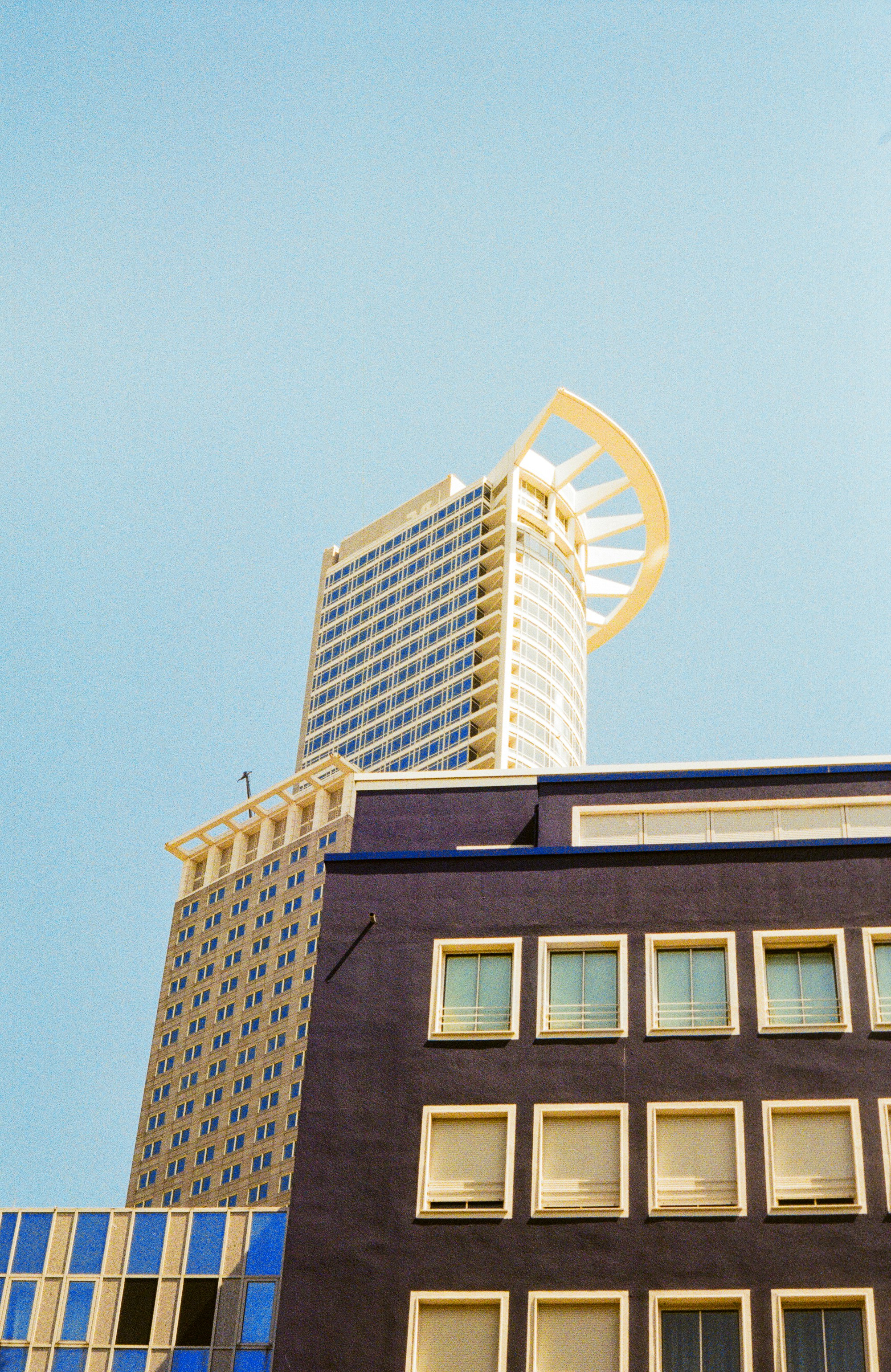 A high-rise building features a modern architectural design with a unique arched structure at the top, contrasting against a clear blue sky. The upper portion of the building displays a grid of reflective windows and a light-colored facade, while the lower section shows a darker block with smaller windows.