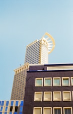 A high-rise building features a modern architectural design with a unique arched structure at the top, contrasting against a clear blue sky. The upper portion of the building displays a grid of reflective windows and a light-colored facade, while the lower section shows a darker block with smaller windows.