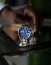 A stylish wristwatch with a blue dial and metallic strap is prominently displayed. It features multiple sub-dials and a date window. The reflection of the watch is visible on a glass surface. A person’s hand is in the background, slightly out of focus.