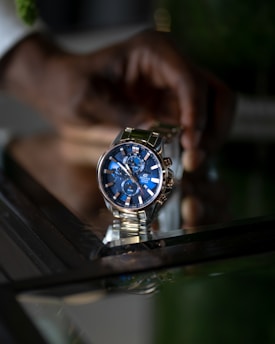 A stylish wristwatch with a blue dial and metallic strap is prominently displayed. It features multiple sub-dials and a date window. The reflection of the watch is visible on a glass surface. A person’s hand is in the background, slightly out of focus.