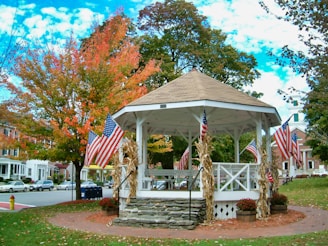 A white gazebo surrounded by trees with autumn foliage is adorned with American flags. Corn stalks are tied to the gazebo's pillars, and potted mums sit at its base. Buildings and parked cars can be seen in the background, typical of a quaint small-town setting.