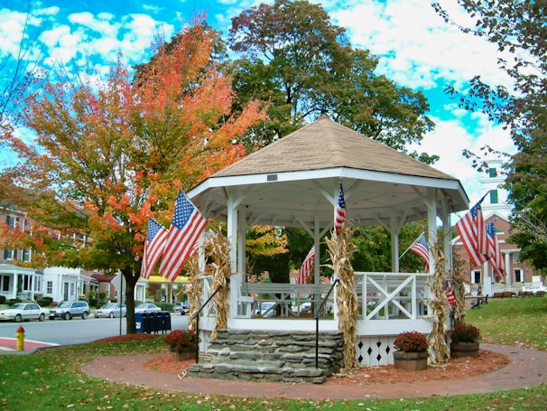 A white gazebo surrounded by trees with autumn foliage is adorned with American flags. Corn stalks are tied to the gazebo's pillars, and potted mums sit at its base. Buildings and parked cars can be seen in the background, typical of a quaint small-town setting.