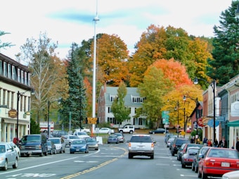 A small town street lined with parked cars on both sides. Several vehicles are driving down the street, and there are people walking along the sidewalks. The buildings have a charming, rustic appearance with some local businesses visible. In the background, there are trees with vibrant fall foliage, displaying hues of orange, red, and green.