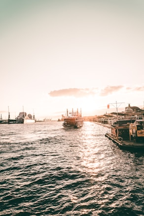 A serene waterfront scene with the sun setting in the background, casting a warm glow over the water. Ships and boats are docked along the shore, with one large vessel prominently featured near the center. The skyline features cranes and buildings, adding an industrial yet scenic backdrop.