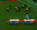Cricketers in blue uniforms walk through a tunnel of fire on a red carpet on a cricket field. They are accompanied by drummers on the left playing large drums, with five women drummers dressed in black. A photographer is seen capturing the moment.