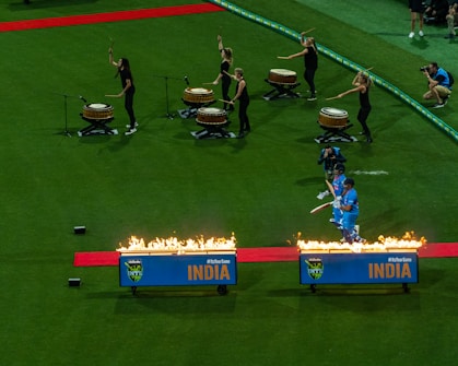 Cricketers in blue uniforms walk through a tunnel of fire on a red carpet on a cricket field. They are accompanied by drummers on the left playing large drums, with five women drummers dressed in black. A photographer is seen capturing the moment.