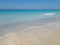 A tranquil beach scene with soft, beige sand leading into clear, turquoise waters. Gentle waves are lapping against the shore under a clear, blue sky.